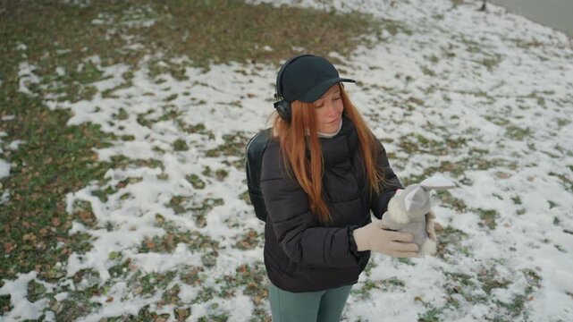 White woman reclaiming snatched plush outdoors wearing headphones and cap extending arms to release toy over snowpatched grass candid upward motion shows relief protective gesture and playful moment