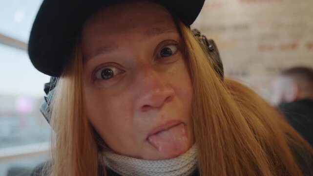 Closeup redhaired white woman wearing headphones, smiling and making playful faces in cozy cafe seat visible scarf and cap, freckles, natural expression, laughter shifting to shy cover of mouth,