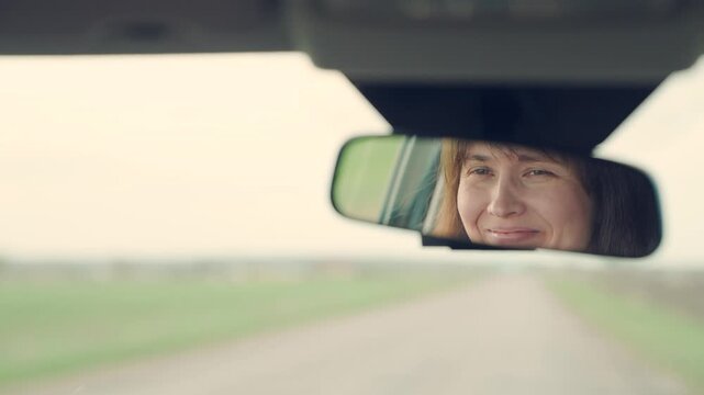 Happy female driver smiles in rearview mirror. Car, road, woman's smile, weekend. Woman driver driving, happy emotions. Successful young woman traveling car, happy smiling, good mood. Trip out of town