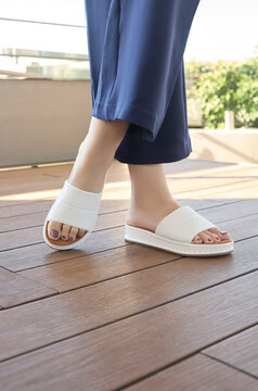 Legs of a young woman modeling stylish white casual sandals, outdoor terrace with soft daylight, wearing navy blue trousers, fair skin, wooden flooring, relaxed style of the footwear, outdoor setting.