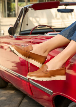 Stylish young woman&rsquo;s legs wearing tan leather loafers with thick soles, with blue jeans, casually resting on a classic red convertible car, outdoors, showcasing a chic, comfortable, casual style.