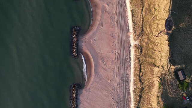 Top down aerial view of sandy beach with waves and rocky breakwater, coastal landscape