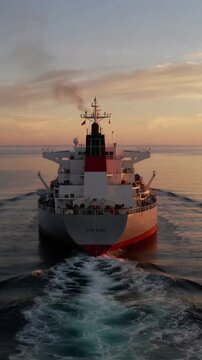 Vertical video: Following drone filming large cargo vessel toward horizon, leaving foamy wake
