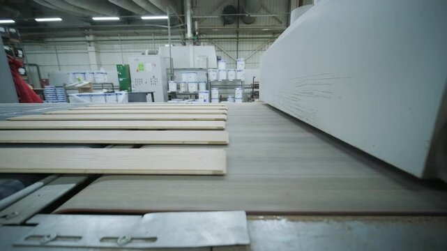 A large wooden panel moves along an industrial blue conveyor belt in a modern woodworking factory. The shot shows safety fences