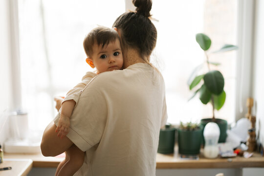 Mother holds baby close in bright kitchen with plants and natural light