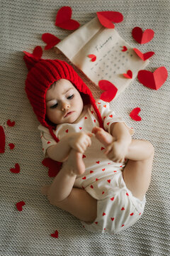 Tiny baby sleeps peacefully in red heart-themed outfit surrounded by scattered paper hearts on soft textured blanket