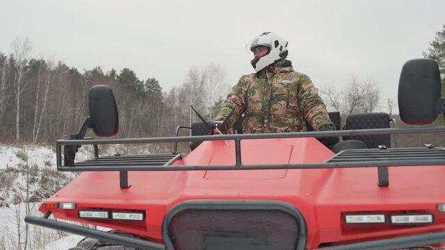 Caucasian rider on red atv in snowy forest, camo jacket and white helmet, closeup front view of vehicle, snowcovered tires, rider surveying trail with steady posture, rugged extreme outdoor mood