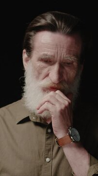 Vertical view of mature Caucasian man touching beard and thinking. Male covering mouth with hand and looking down. Reflecting on memories and past. Standing in dark background in studio.