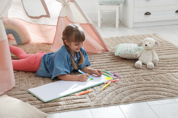 Little girl drawing in sketchbook near toy wigwam at home © New Africa