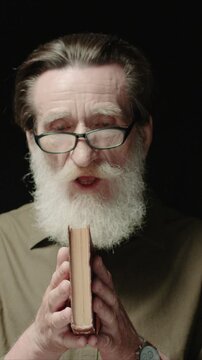Senior Caucasian man wearing glasses blowing dust from closed book. Male holding old book with both hands. Releasing particles into air and watching them. Standing in dark studio background.