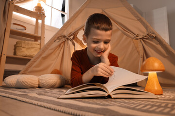 Little boy reading book near toy wigwam at home © New Africa