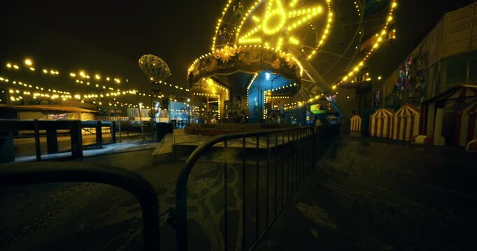 A vibrant carnival is aglow with colorful lights, featuring a carousel and a Ferris wheel. Stalls line the walkway, inviting visitors to enjoy the festive nighttime activities.