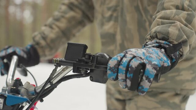 Gloved hands on atv handlebar, camo jacket in snowy forest, adjusting throttle and camera mount, closeup of insulated winter gloves demonstrating secure grip and precise control during extreme trail