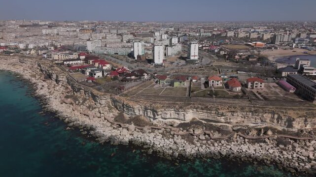 Spectacular aerial drone pull-away shot revealing the Aktau coastline panorama. View of modern residential complexes, private villas, and a developed walking promenade on the rocky Caspian Sea shore. 