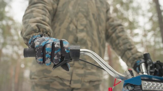 Atv rider snowy pine forest grips throttle, turns on headlight and checks radio while scanning trail for route, camo jacket and helmet visible, blue patterned gloves on chrome handlebar, soft winter