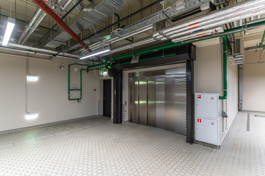 A lift surrounded by exposed pipes and electrical panels. The floor is tiled, and there is bright lighting. The area looks functional and serves as an entry point for the lift