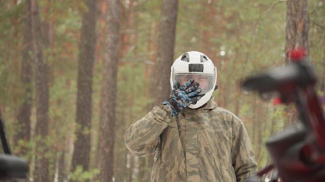 Fullbody rider putting on helmet atv amid pine woodland. Camouflage suit and gloves, white helmet visor and strap secured, rider mounts handlebar and tests control, capturing quiet anticipation