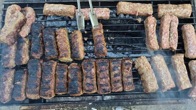 An overhead shot of traditional skinless sausages grilling over hot charcoal. Tongs flip the meat, showing a mix of raw, perfectly browned, and slightly charred pieces on a metal grate.