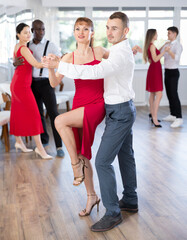 Fototapeta premium Graceful duet, slender woman in vibrant red dress and man in formal wear, engrossed in passionate tango during group dance class in choreographic studio