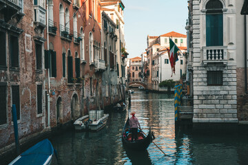 Traditional gondola with a gondolier navigating a scenic Venetian canal. Historic buildings, an Italian flag, and a stone bridge under warm evening sunlight in Venice, Italy. © Maria Sap