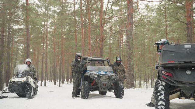 Group riders preparing atv convoy in snowy forest, parked utv and snowmobile line up among tall pines, crew inspects gear and tires before offroad run, calm winter light and crisp air set focused