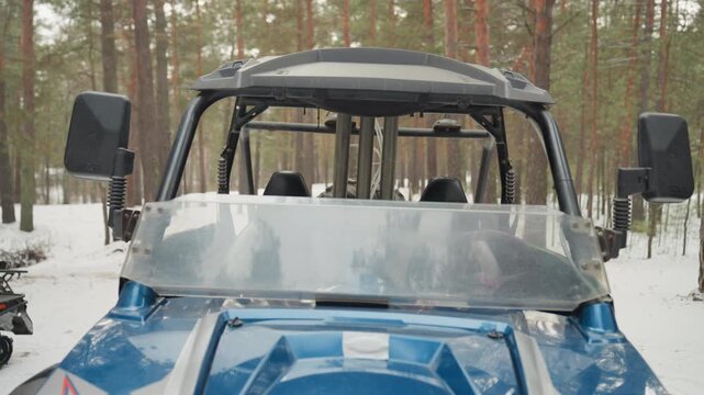 Closeup blue utv with riders preparing two helmeted figures in camouflage and black gear stand beside machine snowmobile and black atv in background winter pine forest frozen ground ready stance