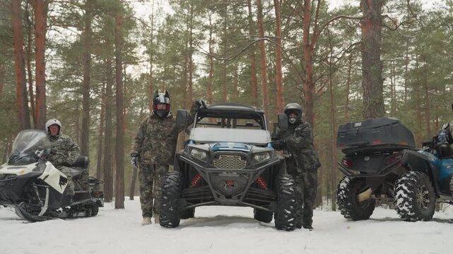 Atv and snowmobile lineup in forest, crew in camouflage gear ready for extreme winter expedition, helmets on, vehicles parked on snow, towering pine trees, falling snowflakes, preride checks and tense