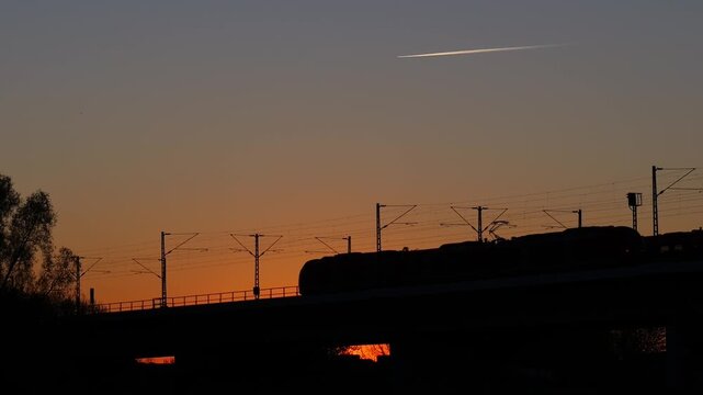 Passenger train silhouette crossing bridge at sunset with orange sky, railway transport scene in Europe