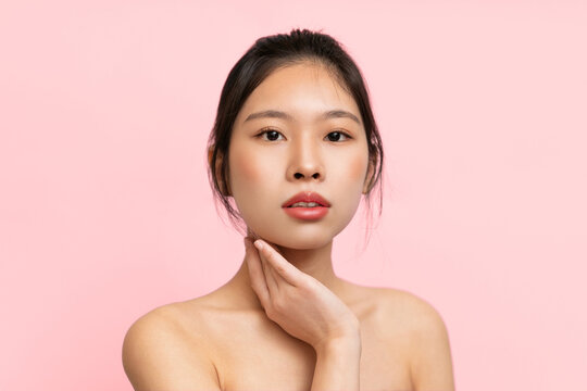 A young model stands in front of a pink backdrop. She poses softy with her hand on her chin. The focus is on her face and makeup during a studio photography session at daytime.