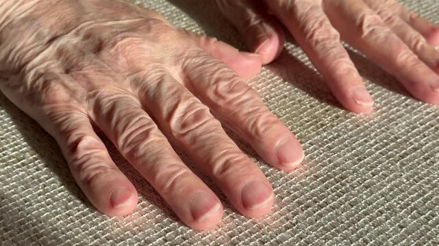 Elderly hands close-up on fabric, sunlight highlighting wrinkles and veins, fingers gently splayed, nails trimmed, soft shadow.