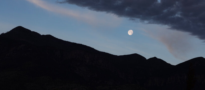 Moon in a blue sky with clouds about to vanish below the Huachuca Mountains in silhouette
