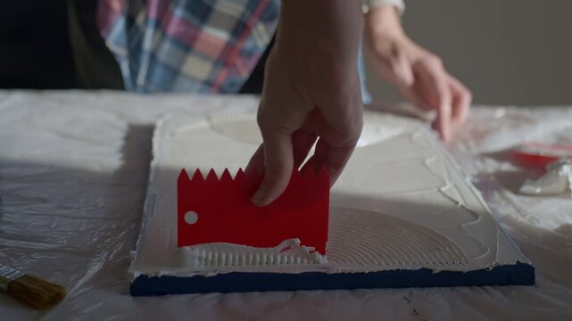 Close up of woman creating textured surface on white canvas using plastic scraper on work table