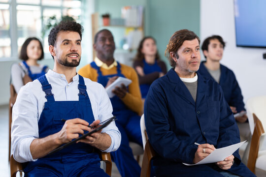 Young man worker in overalls in multinational group of workers listens to lecture and taking notes on tablet in lecture hall