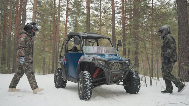 Two riders preparing utv in snowy forest, checking door and gear beside blue sidebyside buggy, helmets on and camo jackets, safety inspection and brief conversation before offroad ride through pine