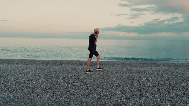 Elderly man walking on the beach near the sea talking on the phone seriously