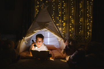 Obraz premium Boy reading book with flashlight in teepee tent at night