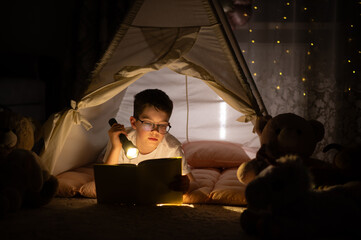 Obraz premium Boy reading book with flashlight in tent at night
