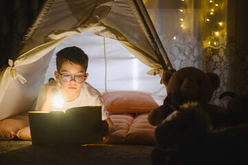 Obraz premium Young boy reading book with flashlight in cozy tent