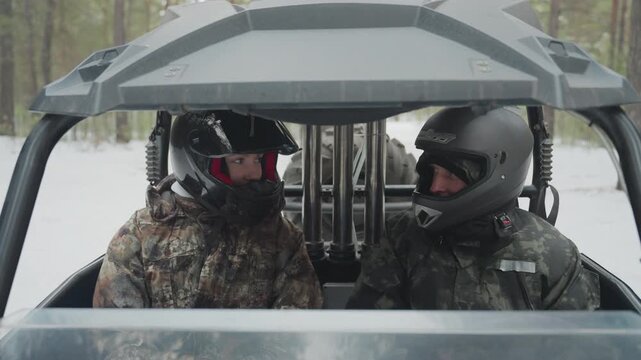 Two white riders in atv utv cabin wearing helmets and camo jackets, seated behind windshield amid snowy pine forest, engines idling, breath visible, tension and camaraderie as they prepare for extreme