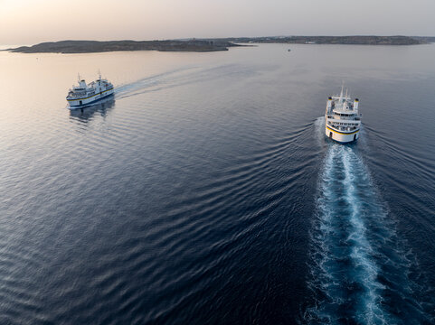 Mgarr Harbor, Malta - 08 June 2025: Aerial view of ferries cutting through the tranquil, deep blue waters towards the distant shore, leaving foamy white trails in their wake.