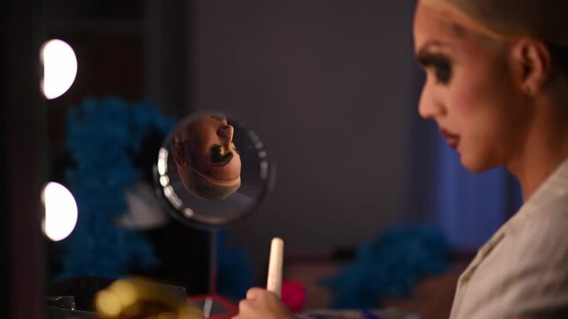Young drag queen carefully applying foundation and contouring makeup with a cosmetic brush in front of a mirror before a show