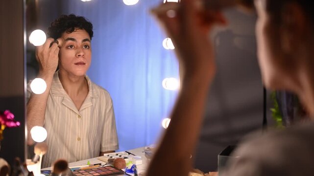 Young drag queen applying foundation and concealer makeup in front of a mirror with lights in a dressing room before a show