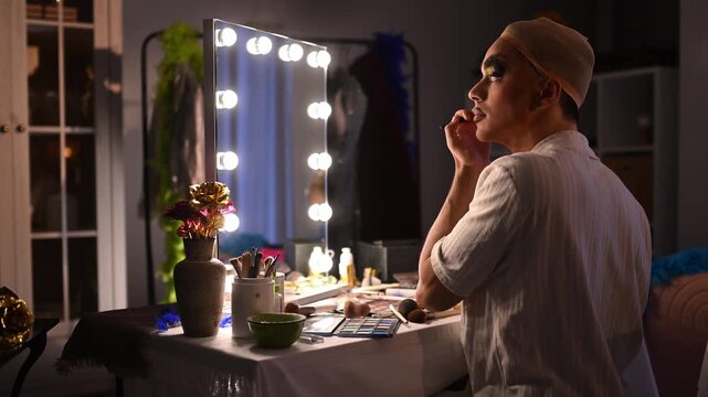 Drag queen applying makeup in front of a vanity mirror while preparing for a show in a dressing room backstage
