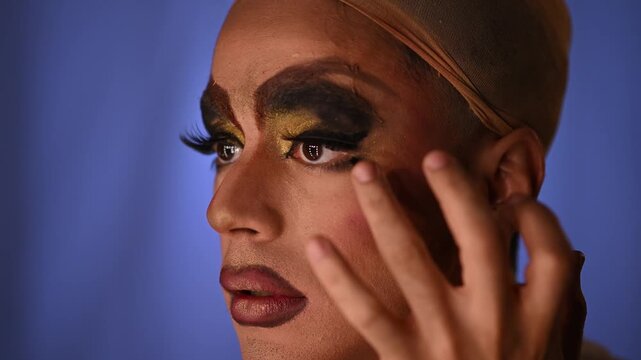 Drag queen applying dramatic eye makeup with false eyelashes and dark lipstick in a dressing room preparing for a show
