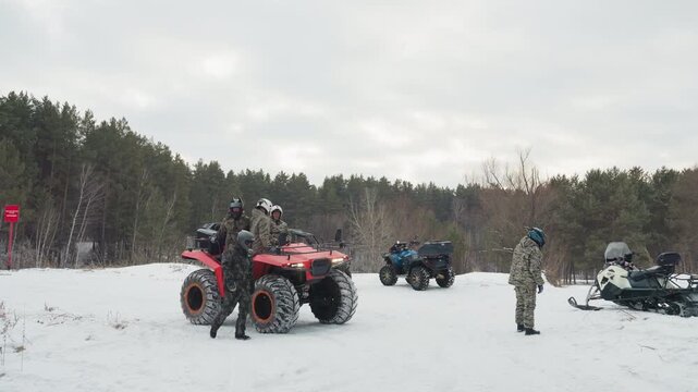 Snowy forest atv meetup with riders preparing machines, red quad in foreground, group checking helmets and gear, snowmobile parked nearby, overcast sky and pine tree backdrop, teamwork and engine
