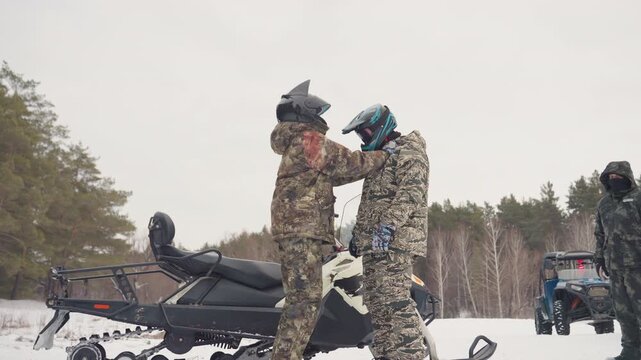 Two riders performing helmet check outdoors beside parked snowmobile, one adjusting visor and strap while crew looks on supportive mentorship, safetyfocused instruction and lastminute readiness create