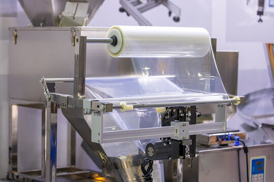 Industrial packaging machine with transparent plastic film roll in a factory setting. Equipment for sealing and wrapping products in manufacturing or food processing industries. Selected focus