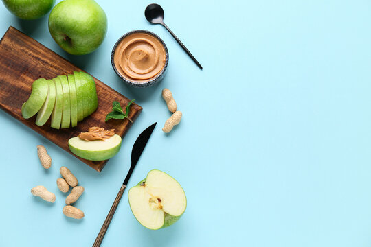 Wooden board with fresh apple slices and bowl of sweet peanut butter on blue background