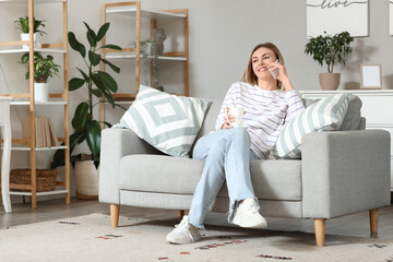 Fototapeta premium Young woman with cup of coffee talking by mobile phone on sofa at home