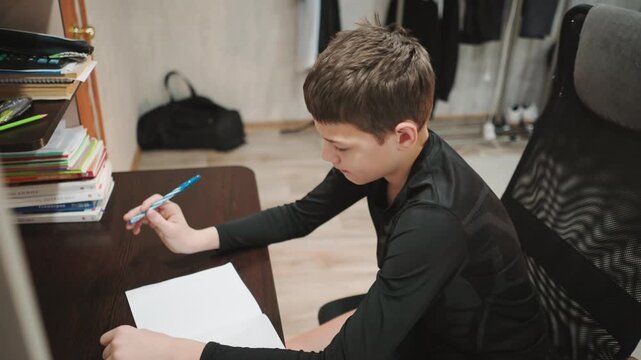 White grandson writing homework at desk, focused expression, leaning over notebook, pen in hand, stack books on shelf, ergonomic chair, hardwood floor, cozy study atmosphere, evening light